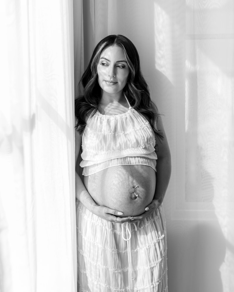 Expectant mother standing in dramatic window light in a black-and-white portrait