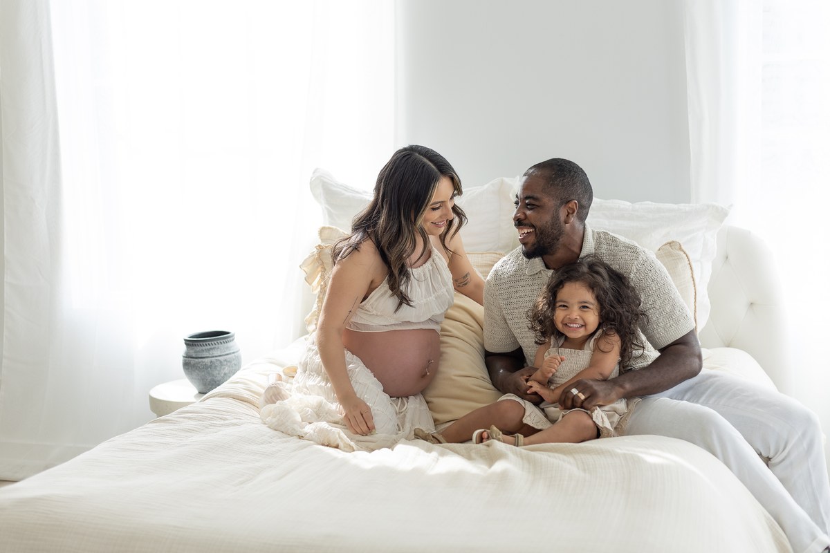 Expectant parents laughing with their young daughter on a sunlit bed
