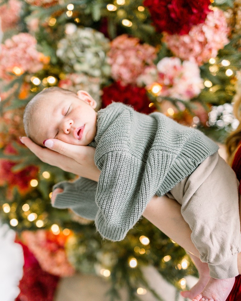Sleeping newborn resting in a parent's hands beside a floral holiday tree
