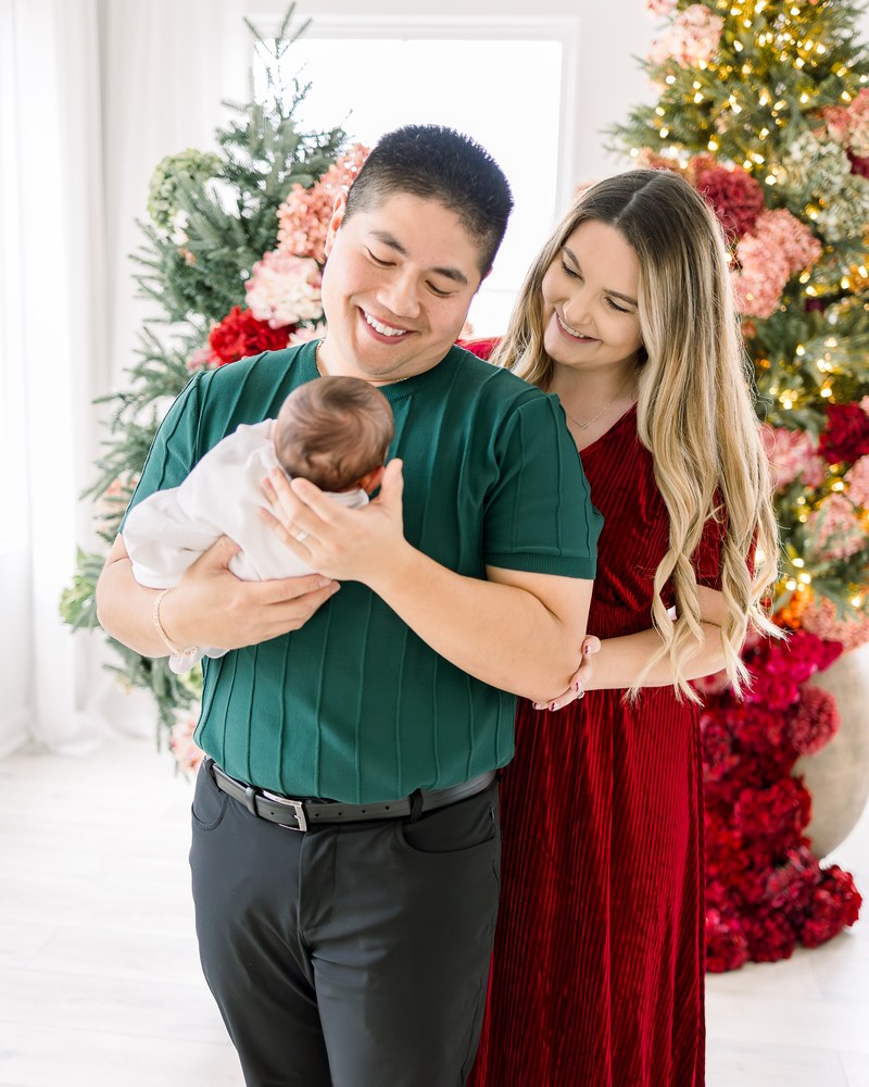 Parents smiling down at their newborn beside a floral holiday tree