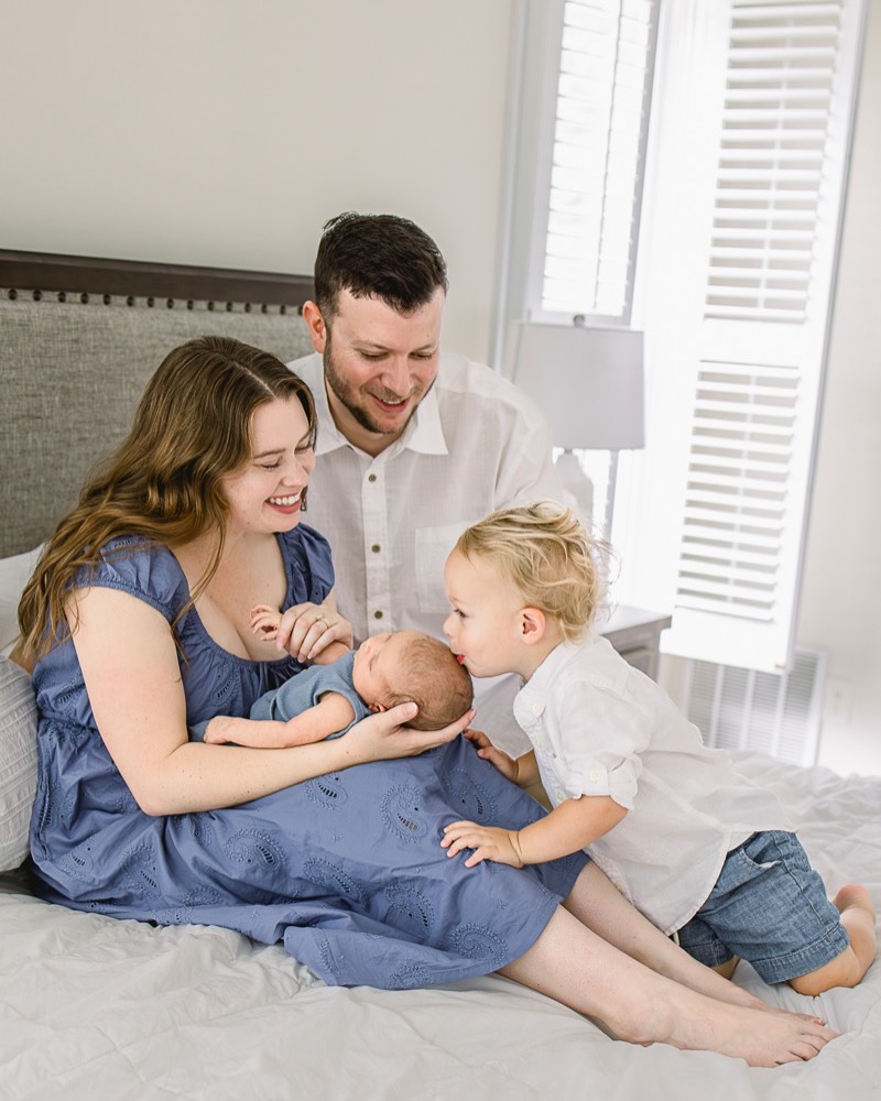 Parents smiling as an older brother kisses their newborn on the bed