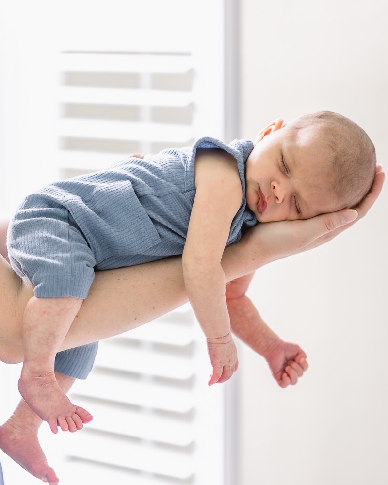 Newborn in a soft blue outfit sleeping across a parent's hands